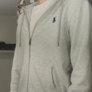 Grå hoodie från Polo Ralph Lauren - Snygg ljusgrå hoodie från Polo Ralph Lauren med dragkedja och huva. Tröjan har två fickor framtill, snörning vid huvan och den klassiska Polo-loggan broderad på bröstet. Perfekt för chill dagar och enkel att matcha med jeans eller joggers.
