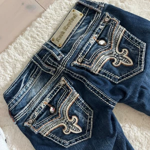 Intressekoll Rock Revival blå bootcut jeans - Intressekoll på ett par snygga Rock Revival jeans i mörkblå tvätt med kontrasterande vita och orangea sömmar. Bootcut-modell med låg midja och detaljerade bakfickor med broderi och blingknappar. Jeansen har klassisk femficksdesign och är gjorda i stretchig denim.