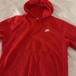 Röd Nike hoodie med huva - Säljer en snygg röd hoodie från Nike med klassisk logga i vitt på bröstet. Tröjan har huva med snörning och en stor magficka framtill. Perfekt för chill dagar eller när du vill ha en sportig look.