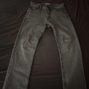 Grå jeans från Jack & Jones - Säljer ett par grå jeans från Jack & Jones med klassisk femficksdesign och raka ben. Jeansen har en snygg tvättad look och är tillverkade i bomull. Perfekta för dig som gillar en enkel och stilren vibe. Storlek 30/30