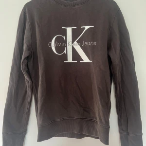 Svart sweatshirt från Calvin Klein Jeans - Säljer en svart sweatshirt från Calvin Klein Jeans med stor vit CK-logga på bröstet. Tröjan har rund hals, långa ärmar och ribbade muddar vid ärmslut och nederkant. Tillverkad i 100% bomull för skön och avslappnad känsla.