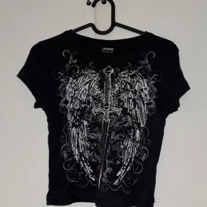 Minga London Svart Baby T-Shirt - Jätte snyg baby t-shirt från Minga London i storlek M (lite åt det mindre hållet) med ett detaljerat tryck av vita vingar och ett svärd på framsidan. Modellen är croppad och har korta ärmar. Använt fåtal gånger, nypris var ungefär 600kr <3