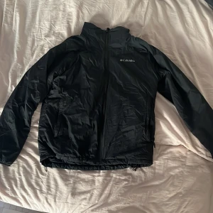 Svart vindjacka från Columbia - Svart vindjacka från Columbia med dragkedja framtill och hög krage. Jackan har två sidofickor med dragkedja och justerbar dragsko nertill. Tillverkad i nylon och elastan, perfekt för blåsiga dagar. Enkel och stilren design med diskret logga på bröstet.