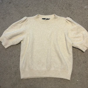 Beige stickad tröja  - Supersöt beige stickad tröja från Vero Moda i storlek S. Tröjan har puffiga korta ärmar och ribbade muddar vid hals, ärmslut och nederkant. Perfekt för dig som gillar en clean och stilren look med lite volym på axlarna.