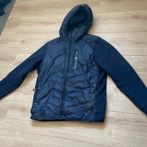 Svart pufferjacka Sail Racing med huva - Snygg mörk blå  pufferjacka från Sail Racing med quiltad framsida och rygg, samt släta ärmar. Jackan har huva, dragkedja framtill och praktiska fickor. Perfekt för kyliga dagar och har en sportig vibe. Materialet är syntet och polyester.