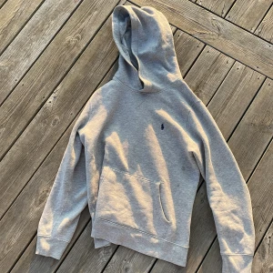 Grå hoodie från Polo Ralph Lauren - Säljer en clean grå hoodie från Polo Ralph Lauren med klassisk broderad logga på bröstet. Tröjan har huva, magficka och långa ärmar. Mjuk och skön bomullsmix.