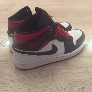 Nike Air Jordan 1 svart/vit/röd - Säljer ett par Nike Air Jordan 1 sneakers i klassisk svart, vit och röd färg. Skorna har röda snören, svart swoosh och perforerad tåbox. Ovandelen är i skinn och de har en ikonisk Jordan-logga på sidan. Perfekta för dig som vill sticka ut med en tidlös streetstyle. Skorna är knappt använda. Öppen för prisförslag 
