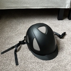 Svart ridhjälm med ventiler - Säljer en svart ridhjälm med flera silvriga ventilationsgaller för extra luftflöde. Hjälmen har justerbart hakband och en snygg, matt finish. Insidan är vadderad för skön passform och extra skydd. Perfekt för dig som vill ha både stil och säkerhet i stallet.