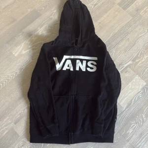 Svart hoodie från Vans med dragkedja - Svart hoodie från Vans i storlek L (barn) med stor vit logga framtill. Tröjan har huva, hel dragkedja och två fickor framtill. Mjuk insida och klassisk streetstil, perfekt för dig som gillar avslappnade vibes.