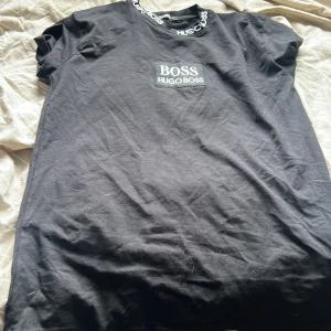Svart t-shirt från Hugo Boss - Svart t-shirt från Hugo Boss med klassisk logga på bröstet och tryckt text runt halsringningen. T-shirten har korta ärmar och är gjord i mjuk bomull. Perfekt för dig som gillar stilrena och snygga basplagg med premiumkänsla.