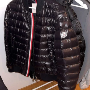 Svart Moncler pufferjacka med dragkedja - Snygg svart pufferjacka från Moncler med glansig finish och klassisk logga på ärmen. Jackan har röd, vit och svart rand vid dragkedjan och ribbade muddar. Perfekt för dig som vill ha en stilren och varm jacka med premiumkänsla.