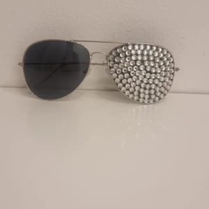 Silvriga aviator-solglasögon med pärlor - Unika aviator-solglasögon med silvrigt metallbåge. Ena linsen är täckt med glittriga pärlor medan den andra är mörkt tonad. Perfekt för dig som vill sticka ut och ha en cool accessoar till din outfit.
