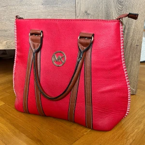 Väska från Michael Kors - Snygg röd axelväska från Michael Kors med bruna handtag och dekorativa sömmar längs sidorna. Väskan har MK-logga i guld framtill och är tillverkad i skinnimitation. Perfekt storlek för att få plats med allt du behöver till vardagen.