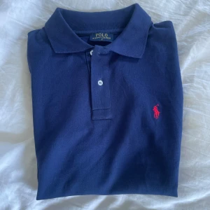 Marinblå pikétröja från Ralph Lauren - Klassisk marinblå pikétröja från Ralph Lauren med röd broderad logga på bröstet. Tröjan är knappt använd och är i storlek S. Skriv för fundering! 