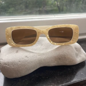 Beige rektangulära solglasögon Gina Tricot - Snygga solglasögon från Gina Tricot med rektangulära bågar i beige, pärlemorskimrande plast. Glasen är bruna och ger UV400-skydd. Perfekt accessoar för att lyfta din stil med en cool retrovibe.