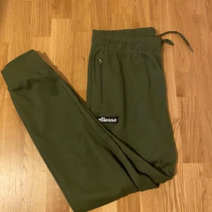Olivgröna mjukisbyxor från Ellesse XL - Snygga olivgröna mjukisbyxor från Ellesse i storlek XL. Byxorna har resår och snörning i midjan, dragkedjeficka och ribbade muddar vid bensluten. Perfekta för chill eller träning, med Ellesse-logga på benet.