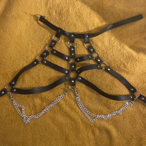 Svart harnessbälte med kedjor - Unikt svart harnessbälte i skinnimitation med flera remmar, silverfärgade nitar och ringar. Utrustad med två avtagbara silverkedjor framtill för extra edge. Perfekt accessoar för att lyfta din outfit och skapa en cool statement-look.