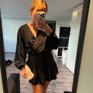 Svart omlottklänning med volang och glitter - Supersnygg svart kort omlottklänning från Gina med subtila glittriga trådar. Klänningen har v-ringning, långa puffärmar och markerad midja med volangkant nertill. Perfekt för dig som vill sticka ut med en elegant men cool look.