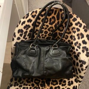 Svart axelväska i skinn med dragkedja - Snygg svart axelväska i skinn med dubbla handtag och guldfärgade metalldetaljer. Väskan har flera fack och dragkedja både fram och baktill. Stilren och rymlig modell som passar till det mesta.