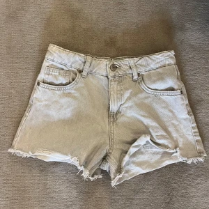 Gråa jeansshorts från Zara - Snygga beige jeansshorts från Zara med hög midja och råa, fransiga benslut. Klassisk femficksmodell med knapp och dragkedja framtill. Perfekta för sommardagar och enkel att matcha med allt.