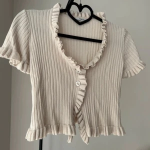 Beige ribbad blus med volang - Supersöt beige ribbad blus från Chiquelle med korta ärmar, volangkant längs halsringning och nederkant samt knappar framtill. Perfekt för dig som gillar romantiska detaljer och en feminin look.