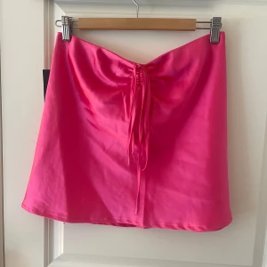 Rosa satintopp från NA-KD - Supersnygg topp i starkt rosa satin från NA-KD. Toppen har en dragsko med snörning framtill som ger en cool detalj och en smickrande passform. Stängs med dragkedja i sidan. Perfekt för dig som vill sticka ut med färg och glans!