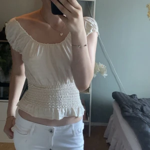 Vit blus med smock från Reflective - Supersöt vit blus från Reflective med smockad midja och volangkant nertill. Blusen har korta puffärmar och en bred, rundad urringning. Perfekt för dig som gillar romantisk och feminin stil. Mjuk och lätt i bomullsmaterial.Det står att storleken är 42 men den är mycket mer som xs eller smal 