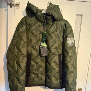 Armani vinterjacka EA7 **superskick** - Snygg grön pufferjacka från EA7 Emporio Armani med quiltad design och  EA7-logga på ärmen. Jackan har huva med smart rotating hood tech och dragkedja framtill. Perfekt för kalla dagar när du vill ha både stil och funktion. Jackan är endast använd ett 10-tal gånger och i mycket bra skick! Lapparna finns kvar enligt bild :) Köpt ny av mig, den har mest hängt i garderoben. 