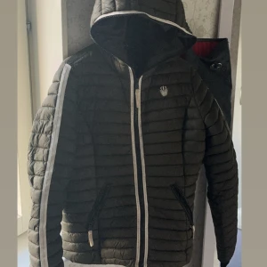 Svart pufferjacka från Navahoo S - Snygg mörk grön  pufferjacka från Navahoo med huva och vita detaljer längs dragkedja och ärmar. Jackan har quiltad design, två fickor med dragkedja och coolt broderat märke på bröstet. Perfekt för kyliga dagar när du vill hålla stilen.