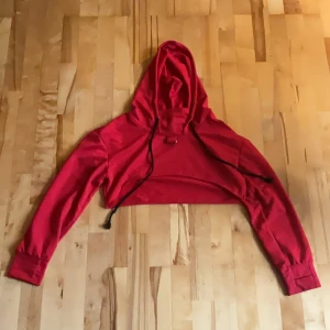 Röd croppad hoodie med huva - Cool röd croppad hoodie med huva och snörning. Tröjan har långa ärmar, breda muddar och en metallring som detalj framtill. Kom egentligen med kedjor som satt fast i ringarna men de har sedan länge sedan gått sönder och är slängda. Tröjan är sitter som en s men armarna kan vara lite korta så passar nog dig som en xs också!
