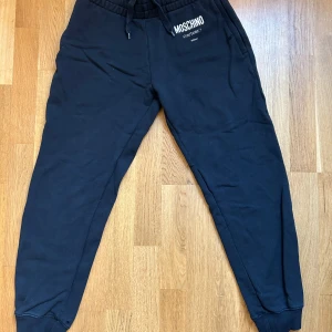 Svarta mjukisbyxor från Moschino - Svarta mjukisbyxor från Moschino Couture med dragsko i midjan och ribbade muddar vid benslut. Byxorna är tillverkade i 100% bomull och har logga tryckt på ena höften. Perfekta för en chill och stilren look.