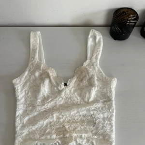 Vit spetsig korsett-topp - Superfin vit korsett-topp med breda axelband och vacker spets över hela plagget. Toppen har en feminin form med markerade kupor och romantiska spetsdetaljer som ger en lyxig känsla. Perfekt att styla med jeans eller kjol för en trendig look.