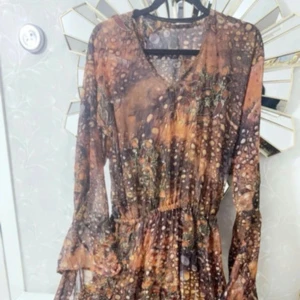 Brun långklänning med mönstrade ärmar - Unik kurdisk långklänning i bruna och orange toner med v-ringning och vida, transparenta ärmar i prickigt mönster. Klänningen har resår i midjan och flowig passform. Materialet är mjukt och glansigt, troligen polyester eller sammet. Perfekt för dig som gillar boho-vibe. Klänningen är justerbar✨
