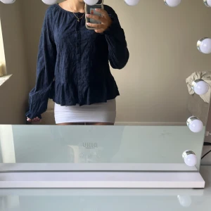 Mörkblå blus med volang och knappar - Supersöt mörkblå blus med volangkant nertill och knappar framtill. Blusen har diskret blommönster i tyget och långa ärmar. Perfekt för dig som gillar en lite romantisk och chill vibe. Sitter löst och har en rund halsringning.