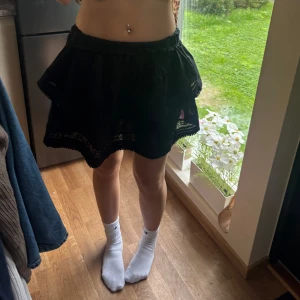 Svart volangkjol med spetsdetaljer - Supersöt svart kortkjol med volanger och fina spetsdetaljer längs nederkanten. Kjolen har en lätt utställd form och resår i midjan för skön passform. Perfekt för dig som gillar en feminin och lekfull stil.