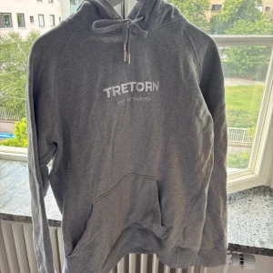 Grå hoodie från Tretorn med tryck - Säljer en grå hoodie från Tretorn med vit logga och text på bröstet. Tröjan har huva med snörning och en stor magficka framtill. Mjuk insida och långa ärmar gör den perfekt för kyligare dagar. Enkel och stilren design som funkar till det mesta.