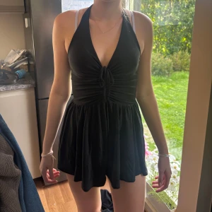 Svart kort halterneck romper - Supersöt svart kort romper med halterneck och djup v-ringning. Klänningen har knytning framtill och ett flowy, utställt kjolparti. Perfekt för varma dagar eller när du vill känna dig extra snygg. Enkel och stilren design som passar till många tillfällen.