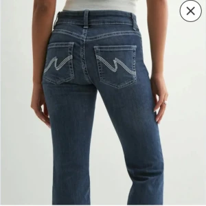 Blå bootcut jeans från Nelly - Snygga mörkblå bootcut jeans från Nelly med hög midja och coola bakfickor med vit zigzag-detalj. Jeansen har dubbla knappar framtill och klassisk femficksdesign. Perfekta för dig som vill ha en tidlös men trendig look. Storlek 34 som motsvarar xs