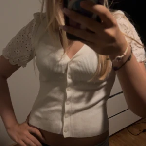 Vit ribbad blus med spetsärmar - Supersöt vit blus med ribbad kropp och korta ärmar i spets med broderade hål och volangkant. Djup v-ringning och knappar framtill.💓