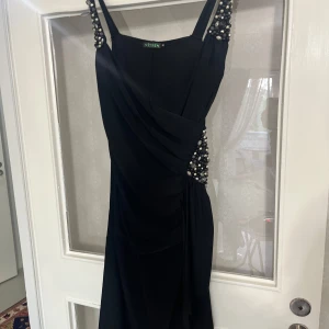 Svart långklänning med pärldetaljer Vitrin - Elegant svart långklänning med draperad design. Klänningen har snygga pärldetaljer på axelbanden och i sidan.