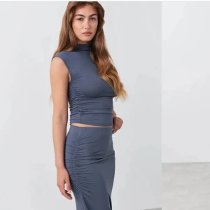 Blå långkjol med en matchande topp - Supersnygg blå långkjol med höghalsad topp och ärmlös design. Setet har rynkade detaljer längs sidorna och en tight passform som framhäver figuren. Perfekt för dig som gillar en stilren och modern look.