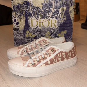 Dior sneakers med monogram och platåsula - Snygga Dior sneakers i rosa och vitt med ikoniskt monogrammönster. Skorna har vit platåsula, rund tå och vita snören med Christian Dior-detalj. Ovandelen är i textil med broderat mönster och insidan har guldfärgad Dior-logga. Perfekta för en trendig look.