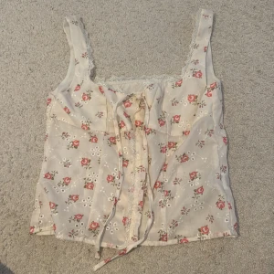 Blommig linnetopp med snörning - Supersöt linnetopp i ljusbeige med små röda blommor och vita broderade detaljer. Toppen har spetskant vid urringningen och snörning framtill för extra detalj. Perfekt för en romantisk och somrig look.