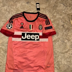 Juventus Dybala #21 rosa Adidas tröja - Juventus fotbollströja med Dybala #21, från Adidas. Rosa med svarta detaljer, vita och röda ränder över bröstet, svarta axelränder och klubbmärke. Tillverkad i lätt och ventilerande Climacool-material. Jeep-logga på magen och Champions League-märken på ärmarna.