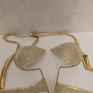 Guldig bikini med strassdetaljer - Supersnygg bikini i guld med glittrande strass över hela tyget. Klassisk trekantsmodell med knyt i nacken och ryggen. Perfekt för dig som vill sticka ut på stranden eller poolpartyt. Materialet är glansigt och känns lyxigt.