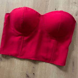 Röd korsettopp från NLY Trend - Snygg röd korsettopp från NLY Trend med vadderade kupor och figurnära passform. Toppen är axelbandslös och har dragkedja i sidan för enkel påklädning. 