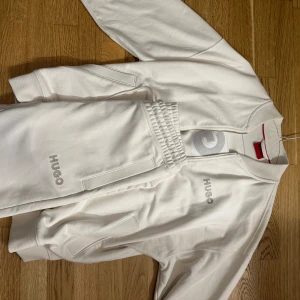 Vit zip sweatshirt från Hugo - Vit sweatshirt från Hugo med half zip och stora logotyper framtill och på ryggen. Tröjan har ribbade muddar, hög krage och är tillverkad i mjuk bomull. Perfekt för en clean och sportig look.