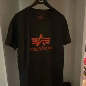Svart Alpha Industries t-shirt - Svart t-shirt från Alpha Industries med orange logga och tryck på bröstet. Klassisk rund halsringning och korta ärmar. Skön passform och tillverkad i mjuk bomull, perfekt för en clean och stilren look. Aldrig använd