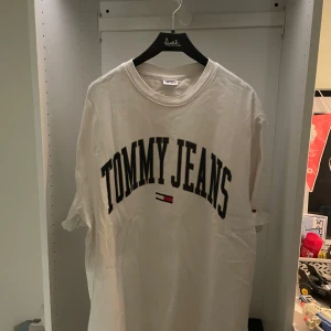 Vit t-shirt Tommy Jeans - Vit t-shirt från Tommy Jeans med stor svart logga över bröstet och klassisk flaggdetalj. Rund halsringning och korta ärmar, perfekt till jeans eller shorts. Oversized. Aldrig använd.