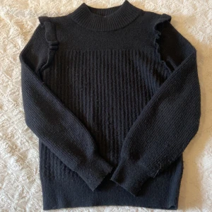 Svart stickad tröja från Stories - Svart stickad tröja från Stories i storlek S. Tröjan har ribbad struktur, högt ribbstickat halslinje och långa ärmar. Snygg volangdetalj på axlarna som ger en trendig touch. Perfekt för lager-på-lager och passar till allt.